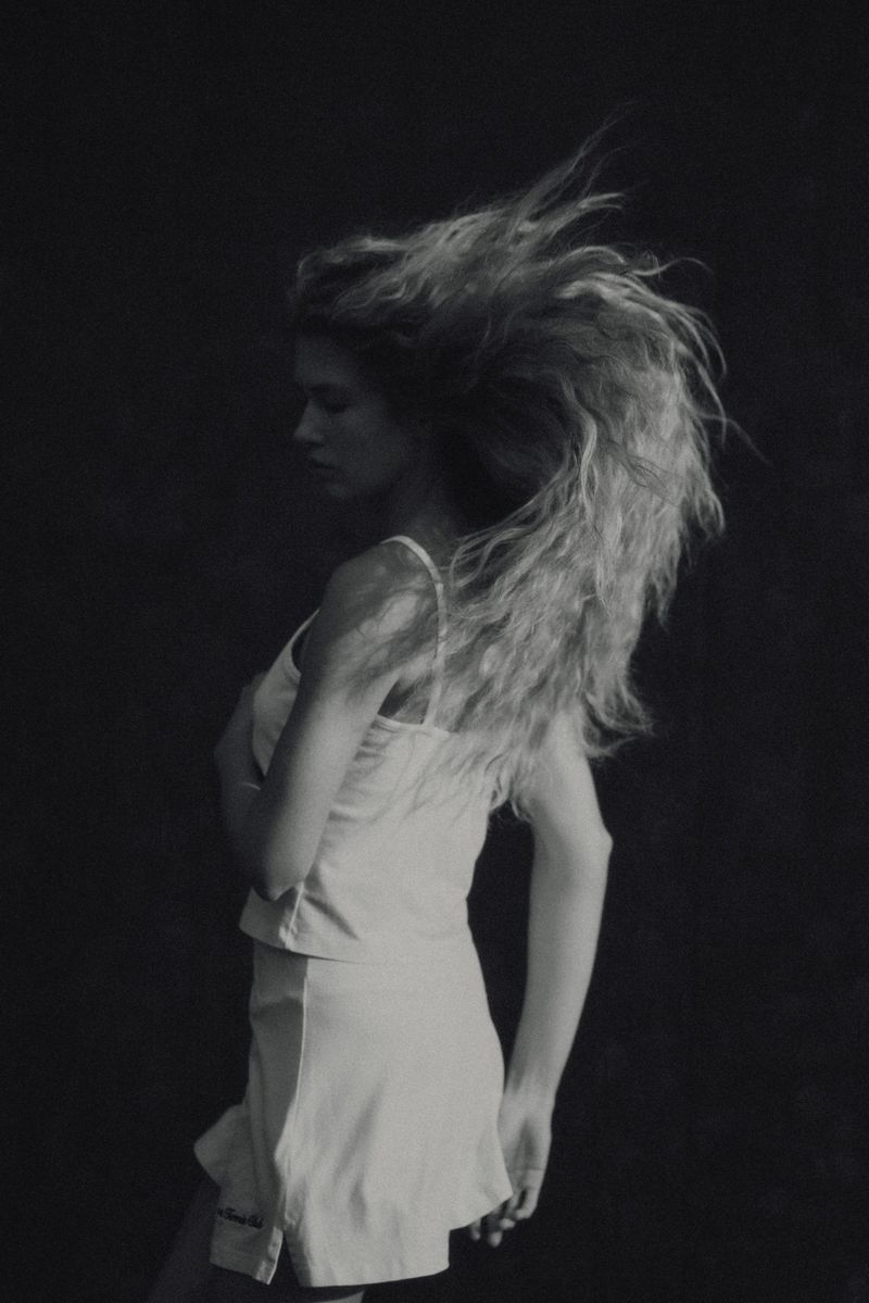 Model Meg Dmitruk with voluminous wavy hair suspended in motion wearing a light tennis top and skirt against a dark background.
