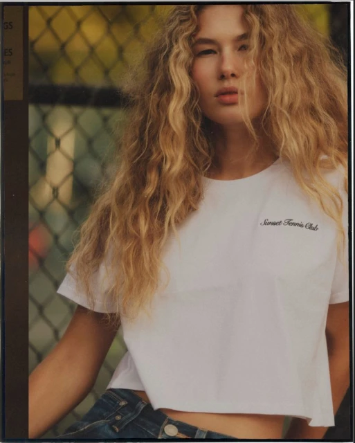 Young woman with curly blonde hair in a white crop top with blue jeans, standing outdoors in front of a chain-link fence and greenery.