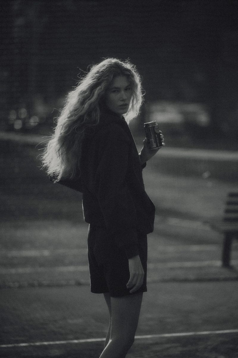 Woman with long, wavy hair stands outdoors holding a can. The grayscale shot features a dark jacket and a park bench in the background.