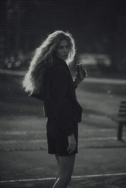 Woman with long, wavy hair stands outdoors holding a can. The grayscale shot features a dark jacket and a park bench in the background.
