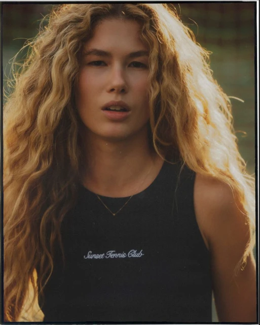 Portrait of a young adult woman with long curly blond hair, wearing a black top, illuminated by natural sunlight.