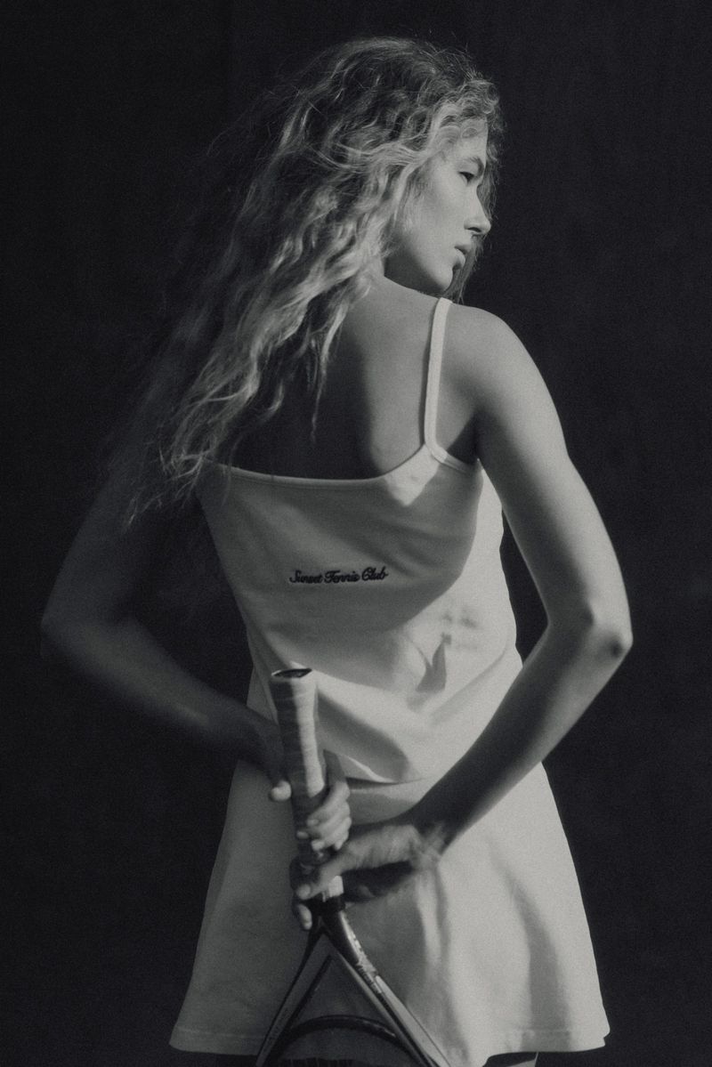 Blonde woman in tennis dress holds a racquet, looking back over her shoulder in a black-and-white studio shot with soft lighting.