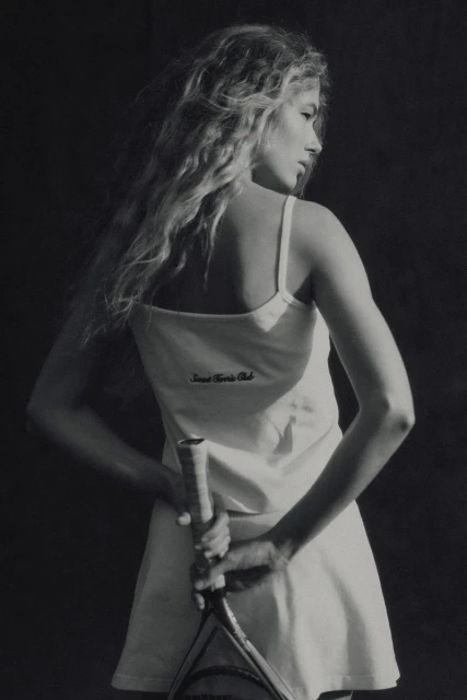 Blonde woman in tennis dress holds a racquet, looking back over her shoulder in a black-and-white studio shot with soft lighting.