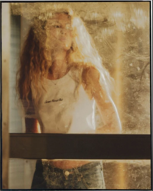 Woman with long blonde hair in a white tank top stands behind a dirty window with a metal frame, looking out into the distance.