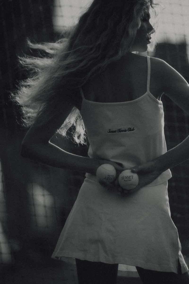 Black-and-white portrait of a woman holding tennis balls, wearing a tennis outfit, standing near a tennis net in an outdoor setting.
