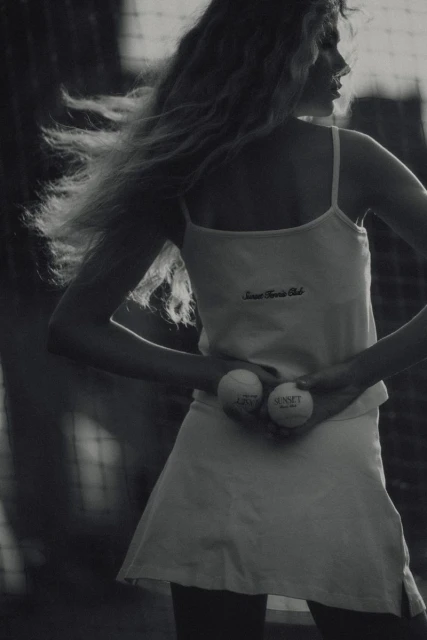Black-and-white portrait of a woman holding tennis balls, wearing a tennis outfit, standing near a tennis net in an outdoor setting.