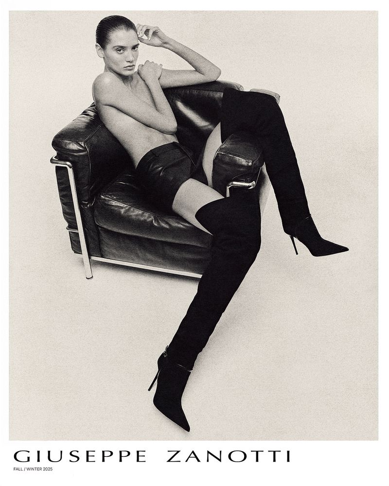 A striking studio shot features a model in high-heeled thigh-high boots, seated in a black chair against an off-white background for fashion.