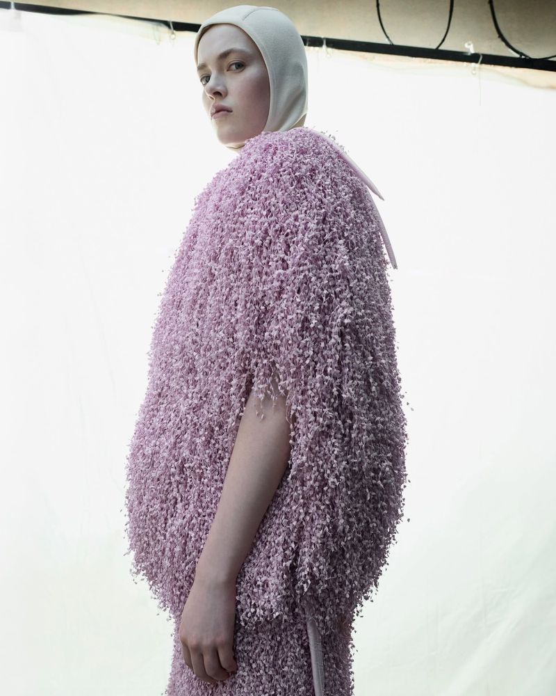 Young woman in textured lavender outerwear and ivory head covering gazes to the side against a bright, neutral background in a fashion shot.