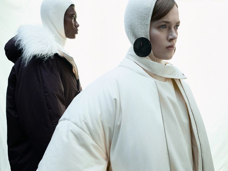 Two women in white balaclavas and jackets, one cream and one dark, stand against a bright white background in a fashion studio shot.