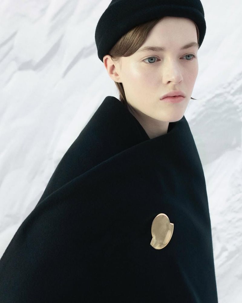 Portrait of a woman wearing a black hat and draped in a black cloth fastened with a large gold brooch against a white background.