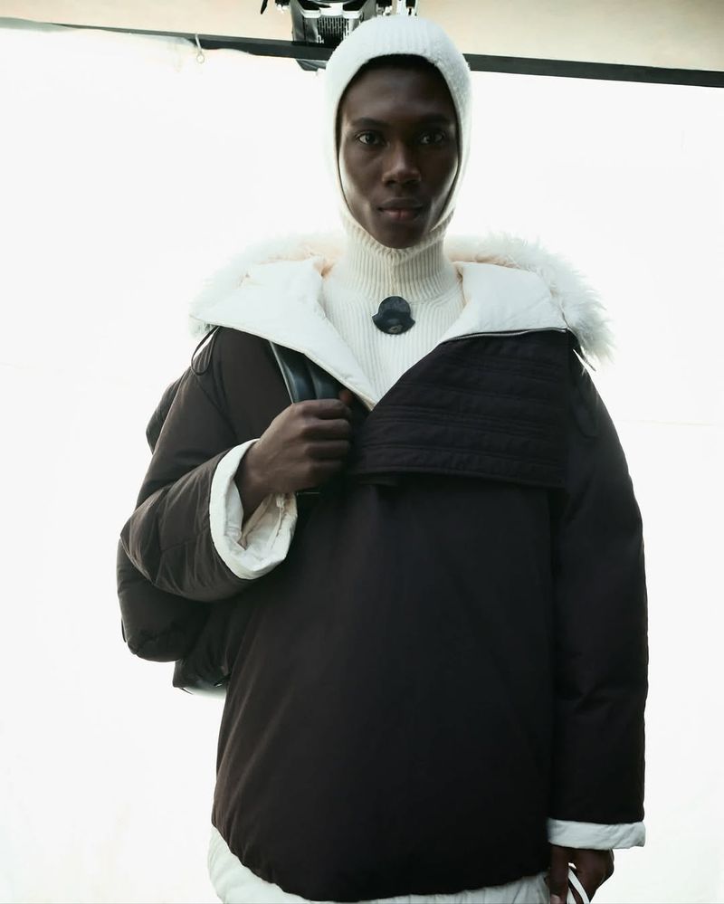 Fashion shot of a Black male model wearing a brown hooded puffer jacket and a white balaclava and turtle neck in a photo studio.