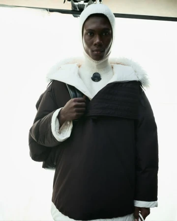 Fashion shot of a Black male model wearing a brown hooded puffer jacket and a white balaclava and turtle neck in a photo studio.