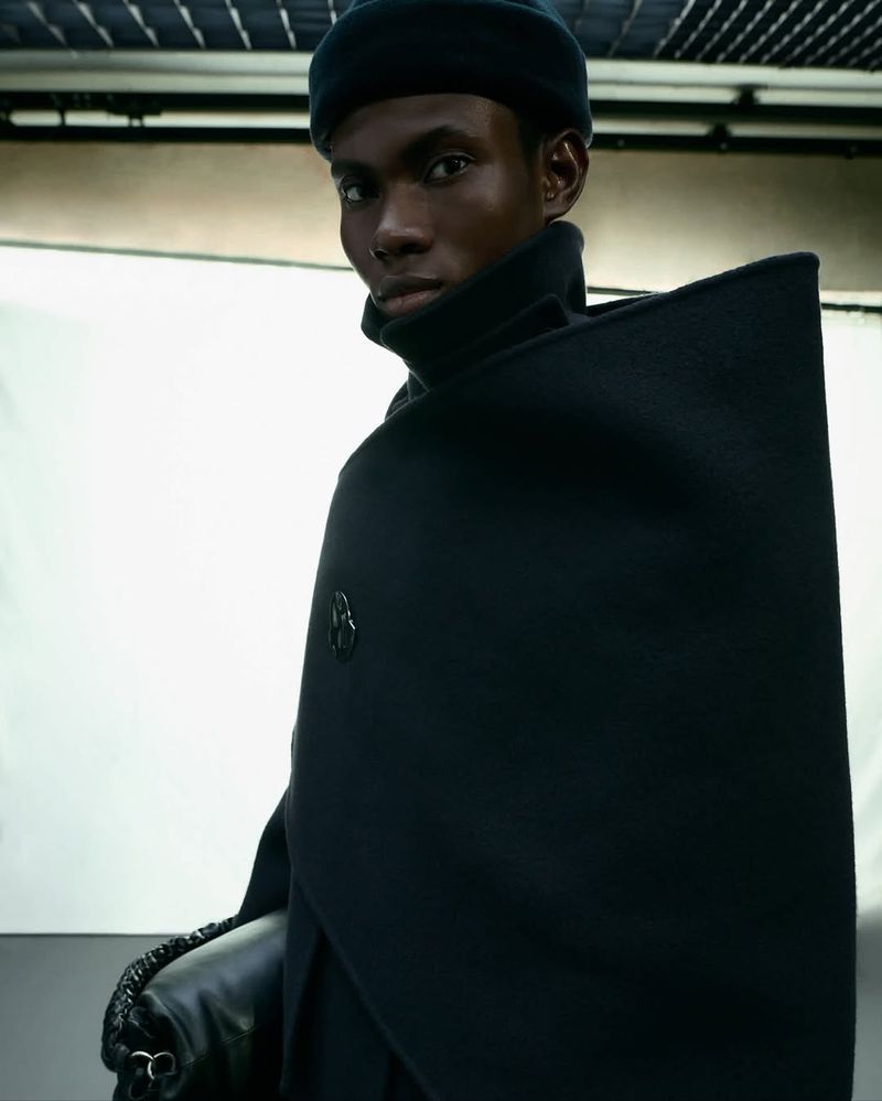 Portrait of a Black male fashion model in black gloves and hat, wearing a dramatic black cape with silver emblem and a high-fashion pose.