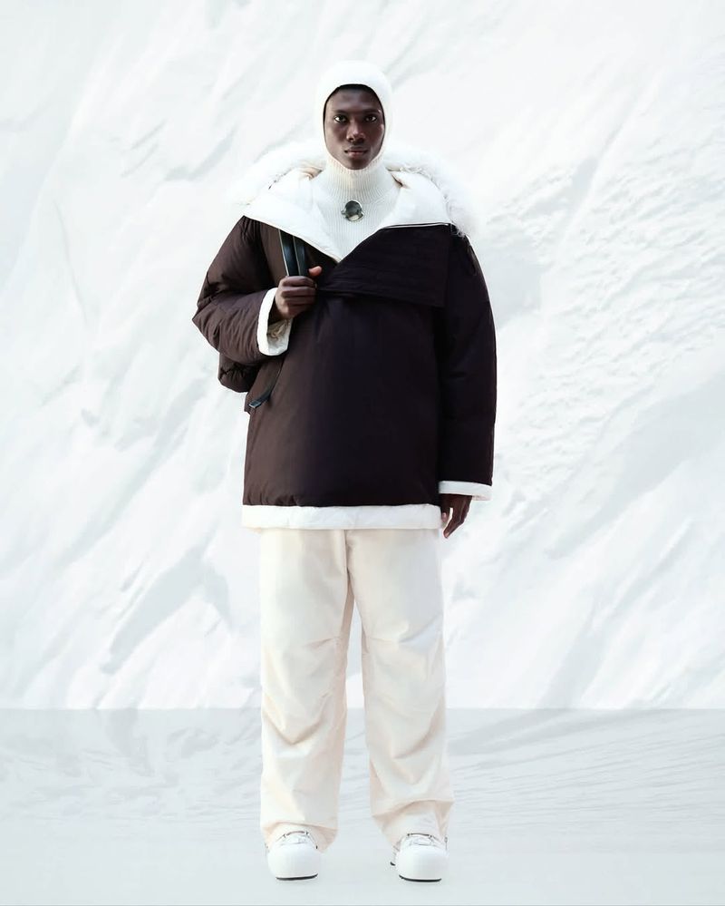 Fashion shot of an adult male wearing a dark brown puffer jacket with white trim, off-white pants, and a white beanie against a white background.