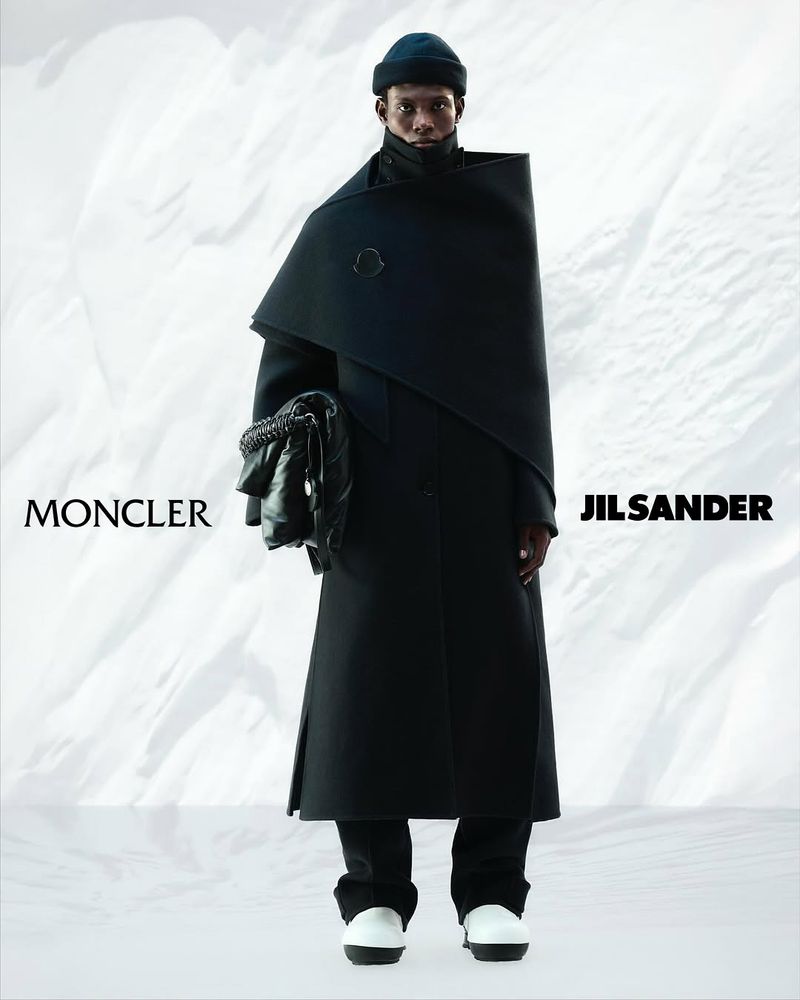 Fashion model in a black coat and beanie, holding a soft black bag against a snowy backdrop, promoting Moncler and Jil Sander.
