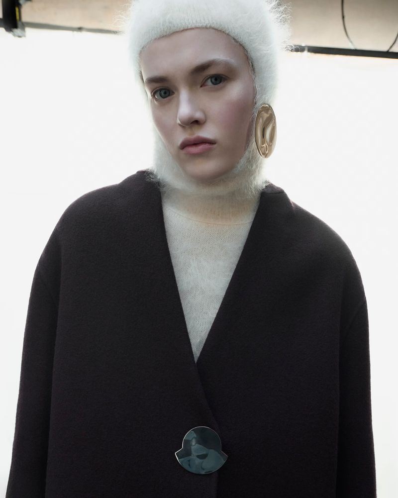 Model Kiki Mckellar wearing a fuzzy white balaclava and an oversized black coat with a logo button against a white backdrop.