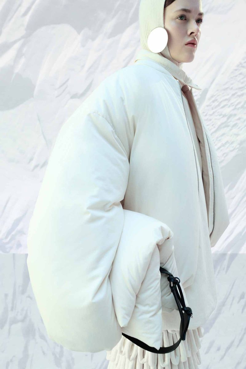 Editorial fashion shot of a woman wearing a large white coat and matching hat against a blurred white abstract background.