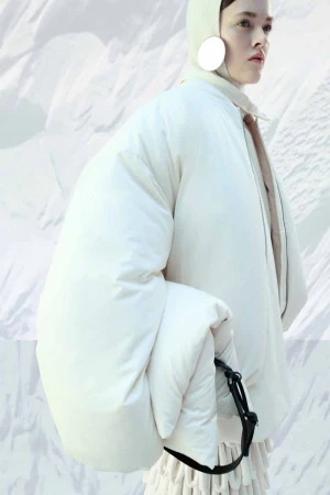 Editorial fashion shot of a woman wearing a large white coat and matching hat against a blurred white abstract background.