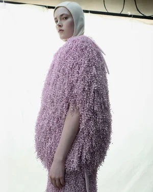 Model wearing a voluminous lilac textured loop-knit cape and cream balaclava against a backlit studio scrim.