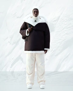 Model wearing a dark brown winter anorak jacket with white trim and cream trousers against a white textured background.