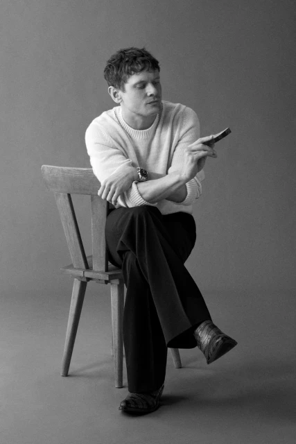 Black and white studio portrait of a fair-skinned adult man seated on a wooden chair, holding a small dark object and glancing down.