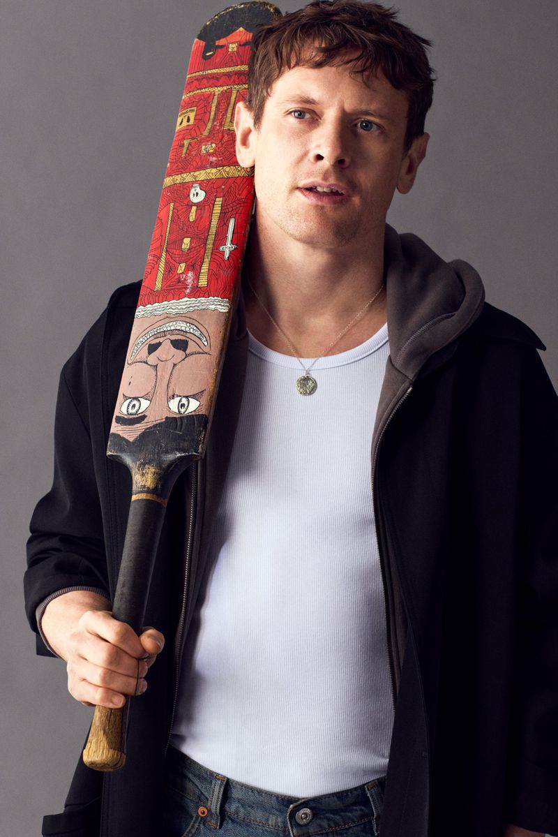 Adult man with painted cricket bat: head and shoulders portrait in front of solid gray background wearing a jacket and jeans.