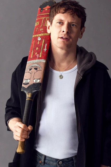 Adult man with painted cricket bat: head and shoulders portrait in front of solid gray background wearing a jacket and jeans.
