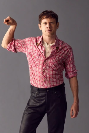 Jack O'Connell posing in a pink plaid shirt and black leather trousers against a grey backdrop.