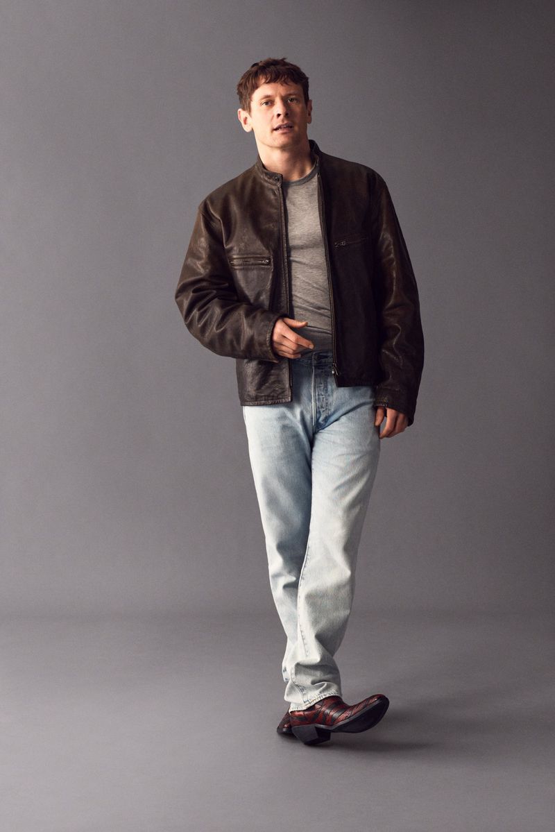Full-length shot of a man in jeans and a brown leather jacket posing in a photo studio against a grey backdrop with diffused lighting.