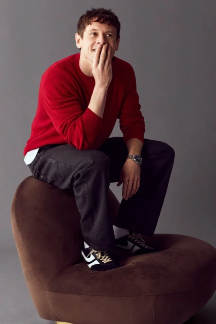 Portrait of a happy adult male in a red sweater sitting on a brown chair with a grey background, showcasing a casual fashion style.