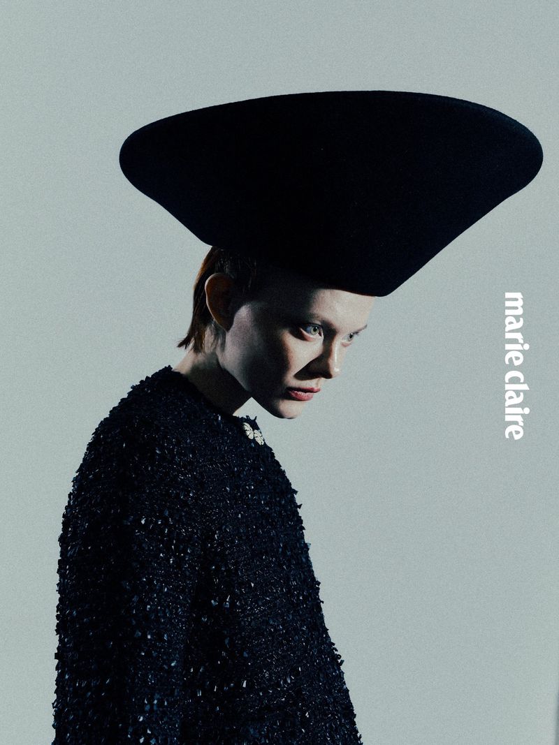 Fashion editorial shot of an adult female wearing a textured black jacket and a large, black hat against a gradient grey background.