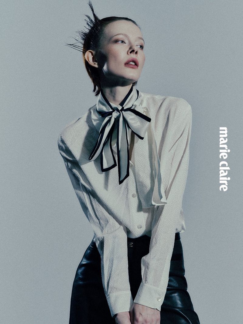 Fashion editorial shot of a young woman with short red hair wearing a cream blouse, black skirt, and striped bow, standing against a gray backdrop.