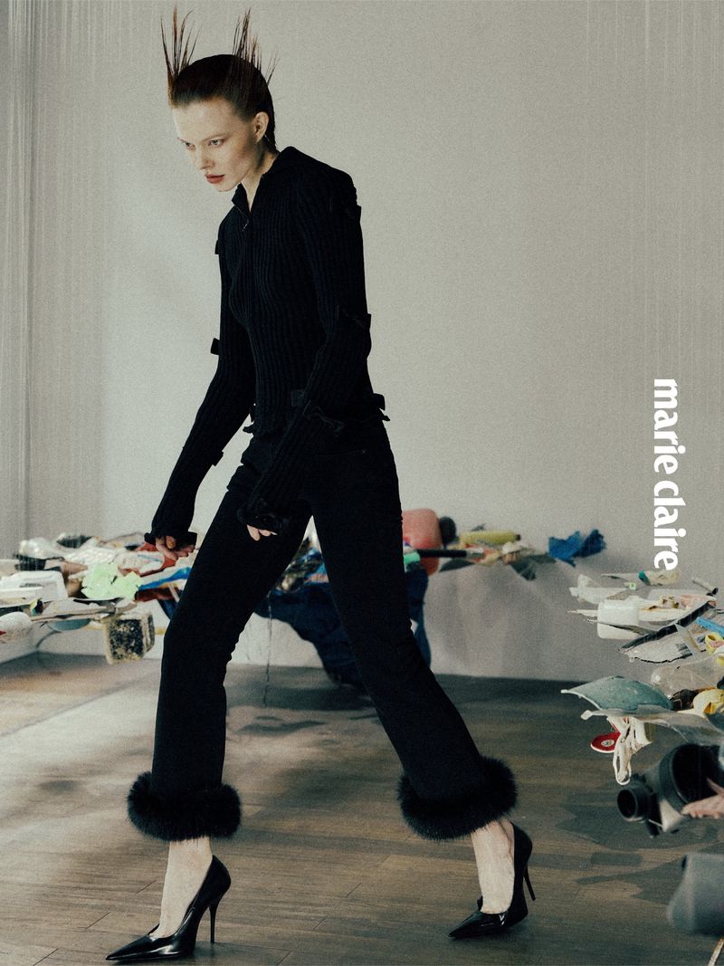 Editorial fashion shot of a woman walking, wearing a black suit with fur trim on the cuffs of the pant legs, posing indoors.
