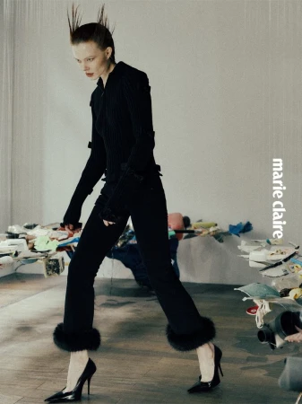 Editorial fashion shot of a woman walking, wearing a black suit with fur trim on the cuffs of the pant legs, posing indoors.