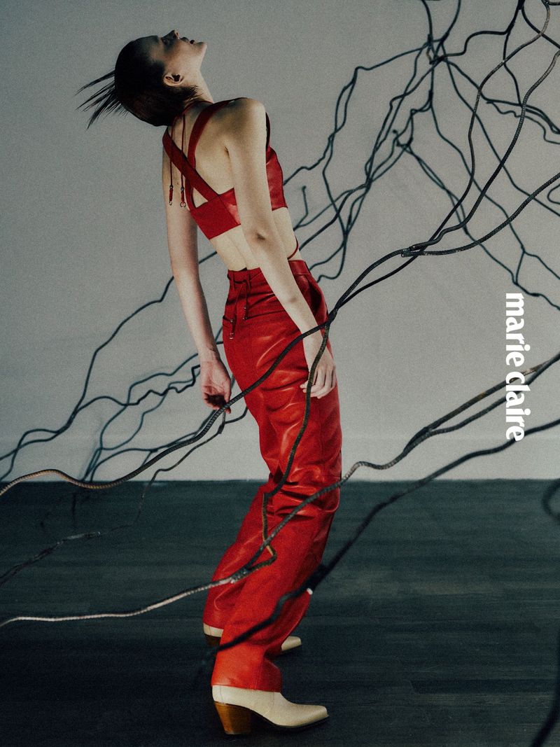 A woman in a red leather outfit poses in a fashion shot against a gray backdrop with black wires. She's wearing heeled boots.