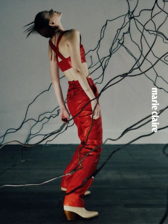 A woman in a red leather outfit poses in a fashion shot against a gray backdrop with black wires. She's wearing heeled boots.