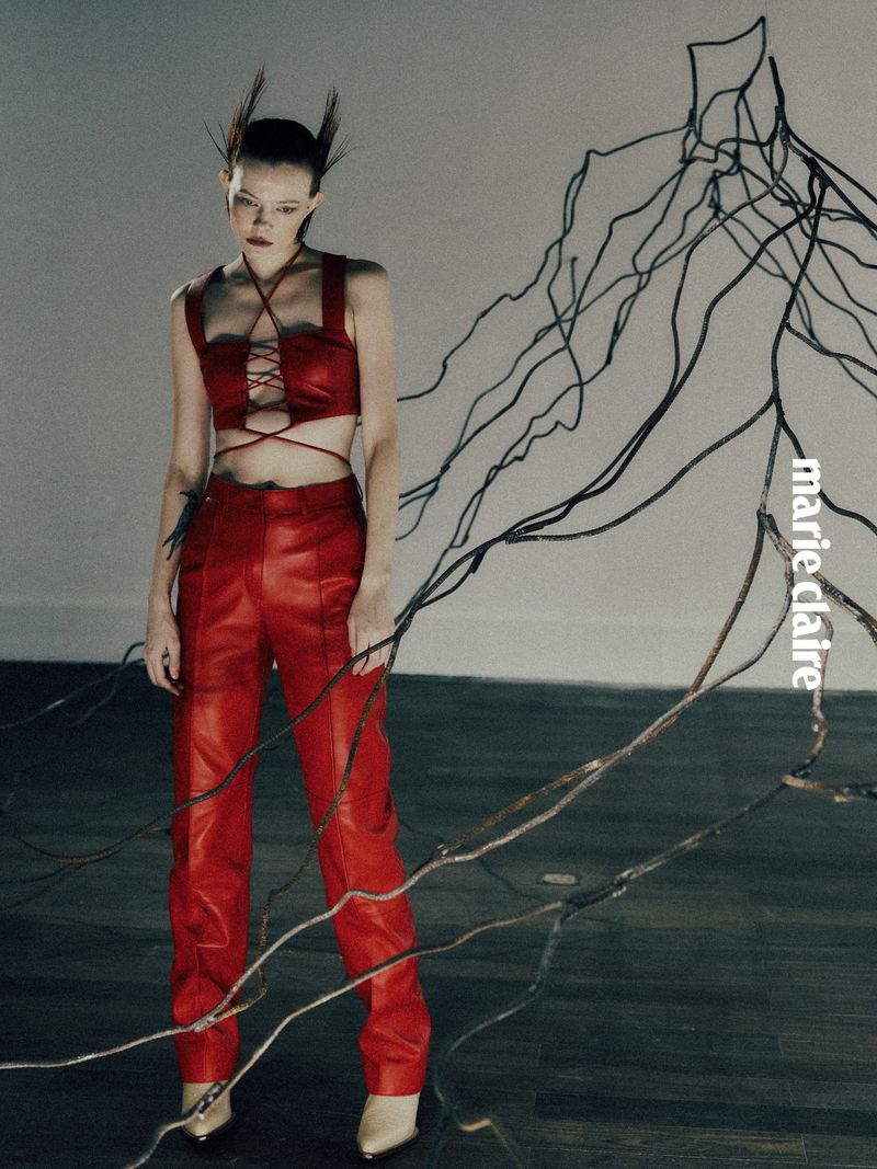A person posing in red top and pants, cream heels, and a unique hairstyle in front of an abstract tree design.