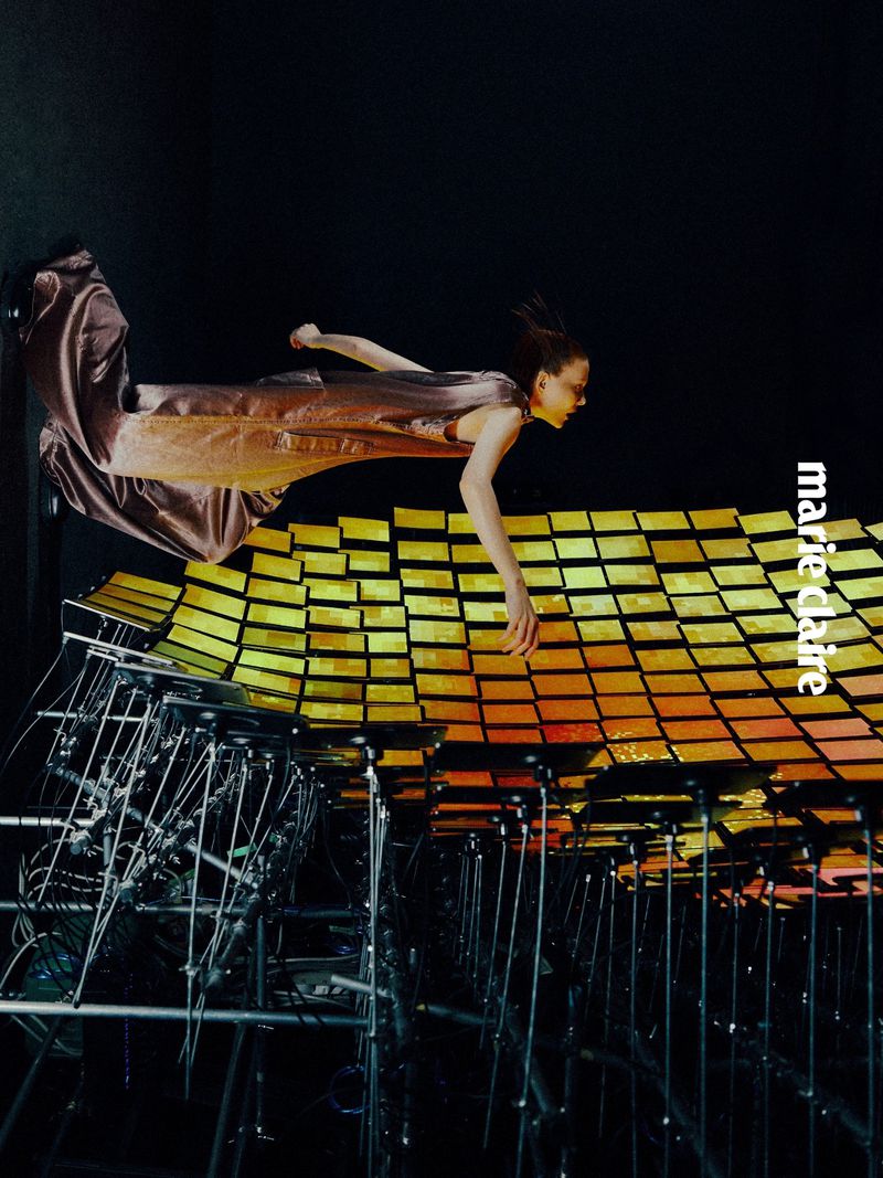 Fashion image: A woman in a flowing gown appears to float above a reflective, gridded surface, captured with an editorial aesthetic against a dark background.