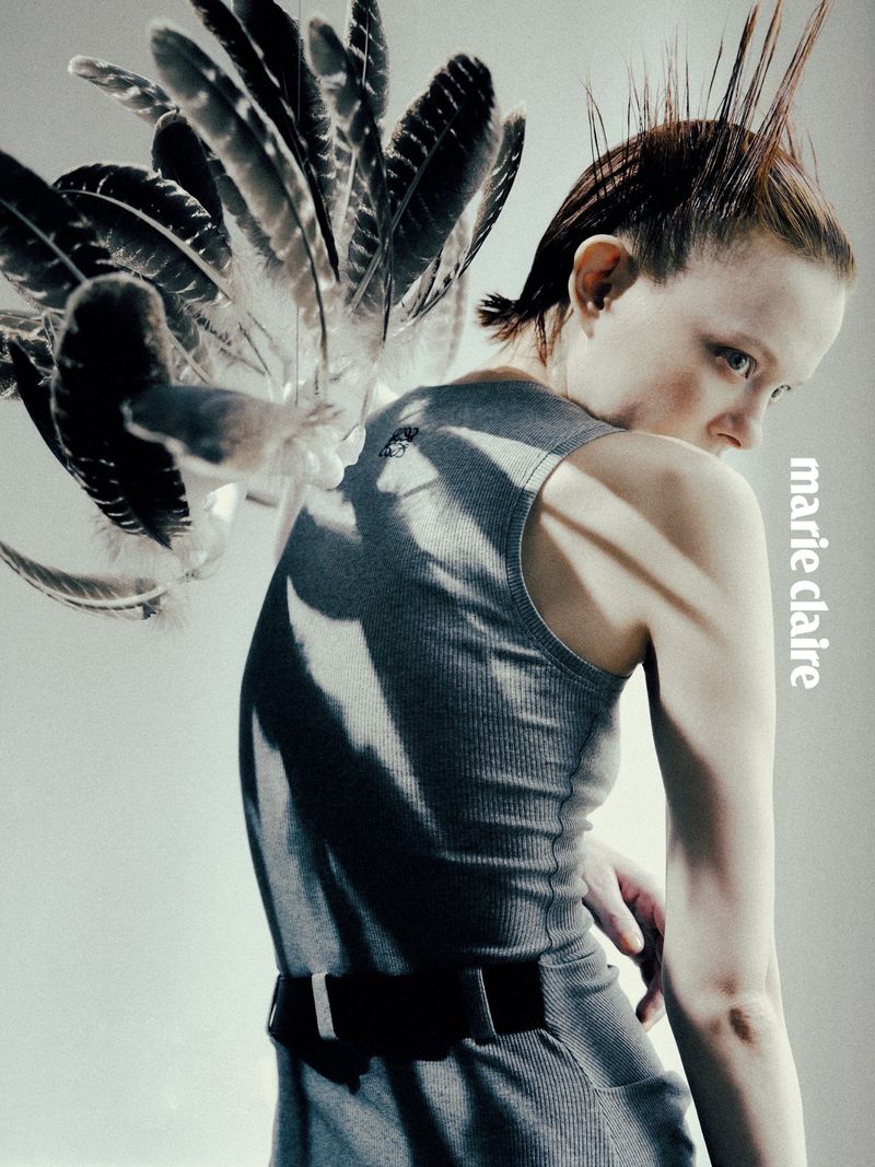 Fashion editorial shot of a woman in a grey dress and spiky hair, feathers accenting her shoulder, looking over her shoulder.