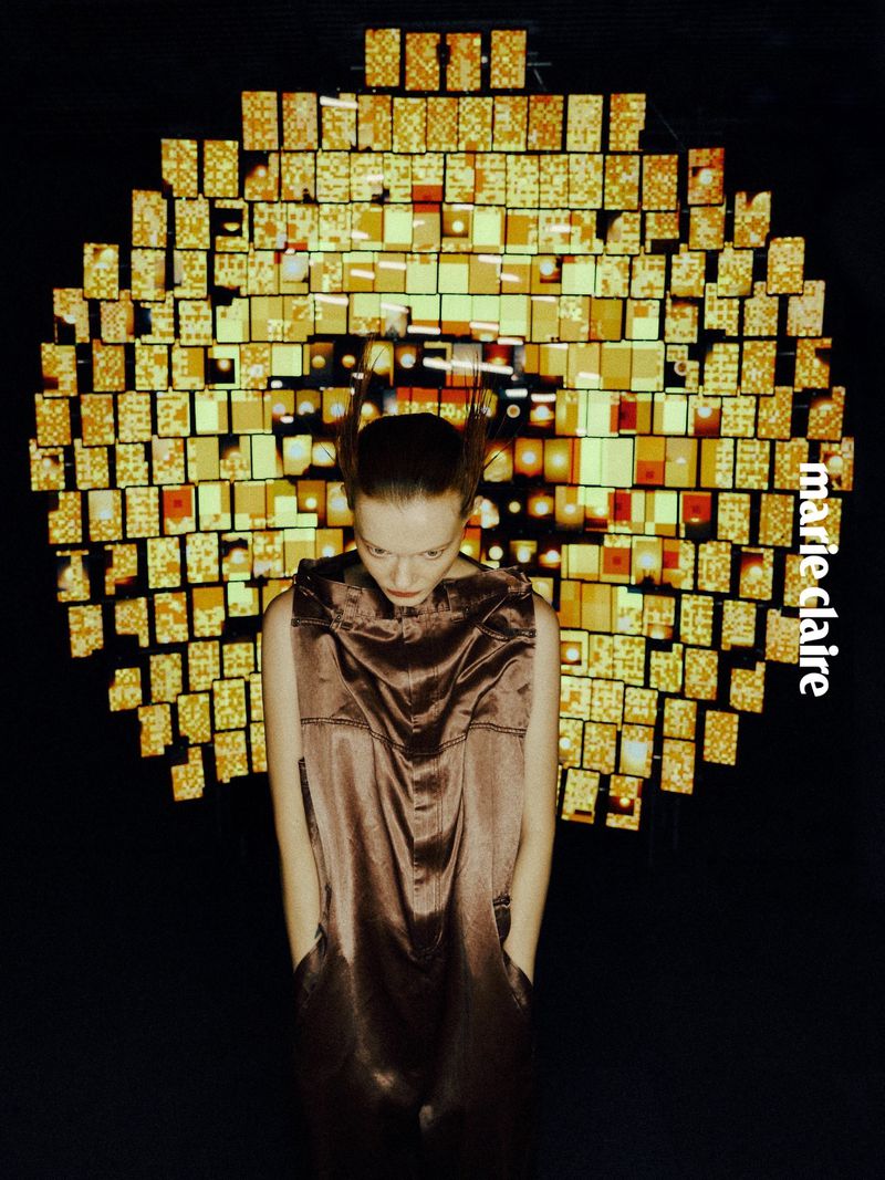 Fashion shot of a young woman in a shiny brown dress looking down, hands in pockets, with a decorative gold background and Marie Claire logo.