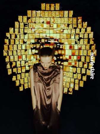 Fashion shot of a young woman in a shiny brown dress looking down, hands in pockets, with a decorative gold background and Marie Claire logo.
