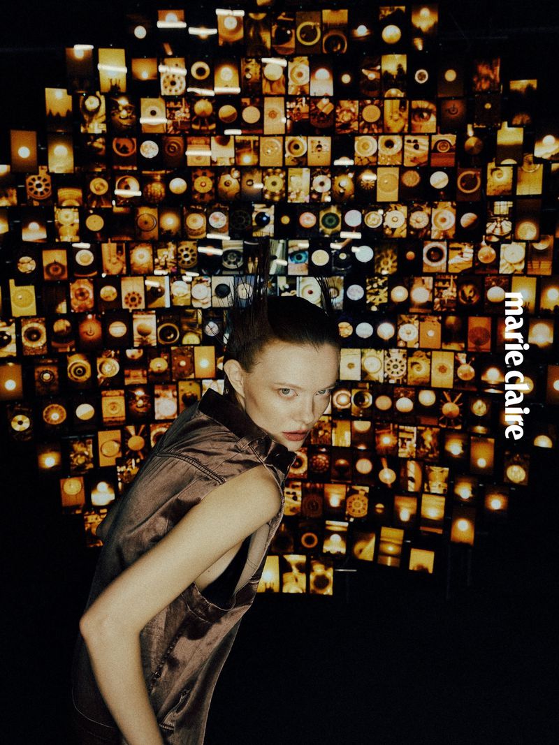 Portrait of a model wearing a sleeveless shirt in front of a wall of vintage cameras. Editorial fashion photography in dark tones.