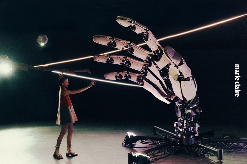 A young woman stands facing a large robotic hand with laser beams in a studio setting, featured in Marie Claire magazine.