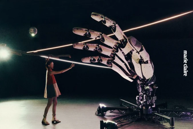 A young woman stands facing a large robotic hand with laser beams in a studio setting, featured in Marie Claire magazine.