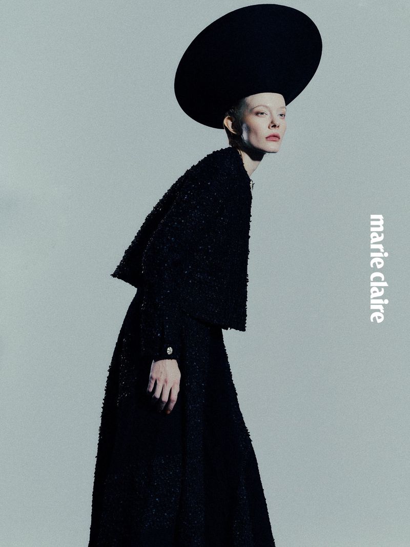 A woman in a black dress and large hat poses in front of a white background for a fashion editorial for Marie Claire magazine.