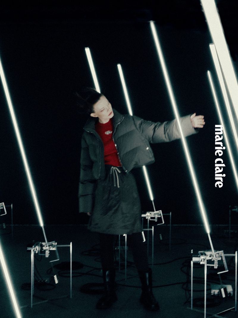 Fashion editorial shot of a model in a puffer jacket and skirt, surrounded by glowing light tubes against a dark background, Marie Claire logo.