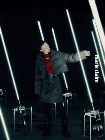 Fashion editorial shot of a model in a puffer jacket and skirt, surrounded by glowing light tubes against a dark background, Marie Claire logo.