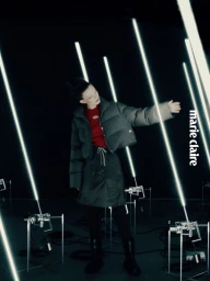 Model Sabi Khazgaleeva wearing a cropped puffer jacket and skirt surrounded by vertical glowing light tubes in a dark studio set.