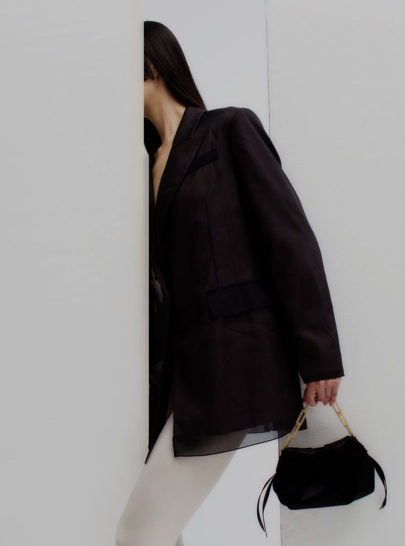 Woman in oversized black blazer with sheer panels and cream pants, holding a black purse with gold chain strap against a white wall.