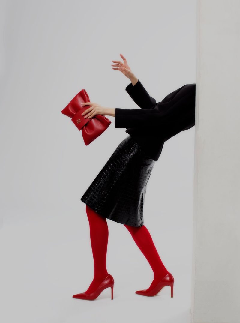 Model strikes a pose with a red handbag in a studio. She wears a black jacket, a matching skirt, red tights and heels, set against a white backdrop.